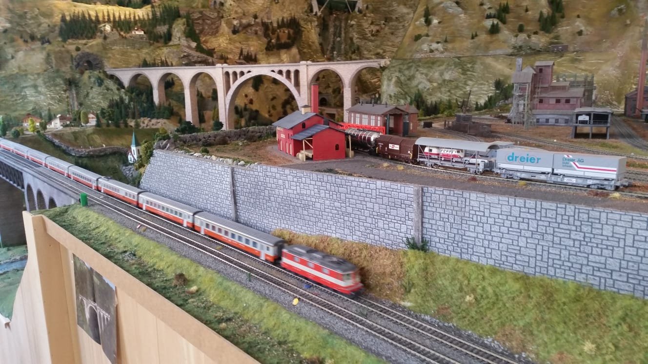 H0-Modellbahnanlage mit Gebirgskulisse, Viadukt und durchfahrendem Zug vor einem Industriegleis